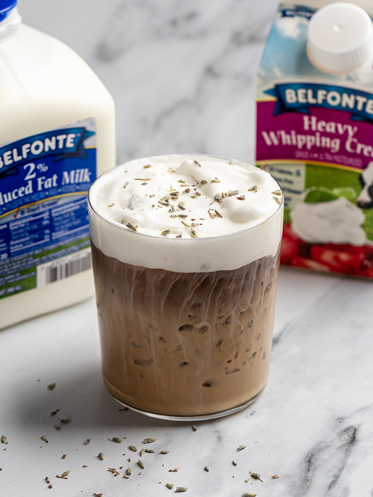 recipe-Honey-Lavender-Cold-Foam-3-post A close-up view of a creamy iced coffee drink topped with frothy cold foam and a garnish of dried lavender, with Belfonte dairy products placed behind it on a marble surface.