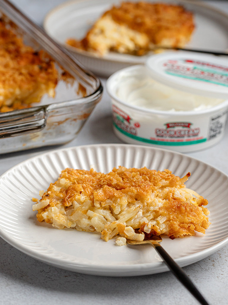 recipe-Shredded-Potato-Casserole-post A serving of shredded potato casserole on a plate with a spoon, with the full baking dish and additional servings in the background along with an open container of Belfonte Sour Cream.