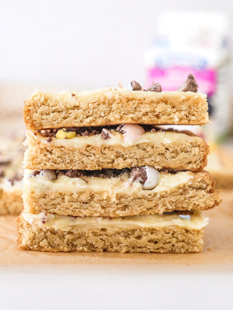 recipe-Sugar-Cookie-Bars-IMG_5645-post A stack of sugar cookie bars with a golden brown base and creamy white frosting, topped with pieces of chocolate candy eggs.