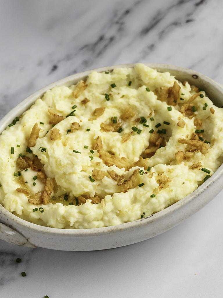 A serving dish filled with fluffy mashed potatoes mixed with French onion dip, garnished with chives and crispy onion pieces, sitting on a smooth marble background.